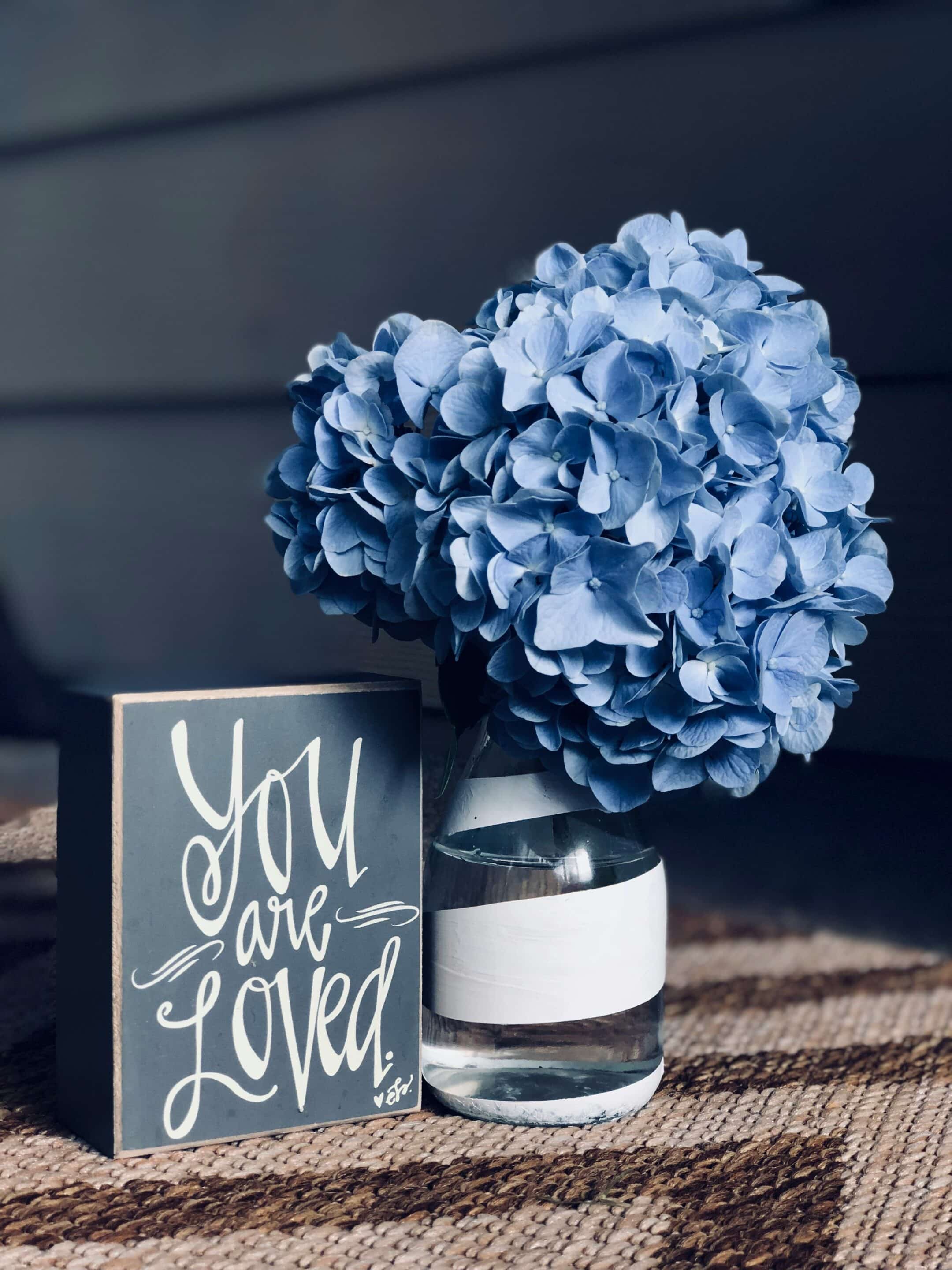 You are loved A chalk board sign which reads you are loved sits next to an arrangement of blue flowers.