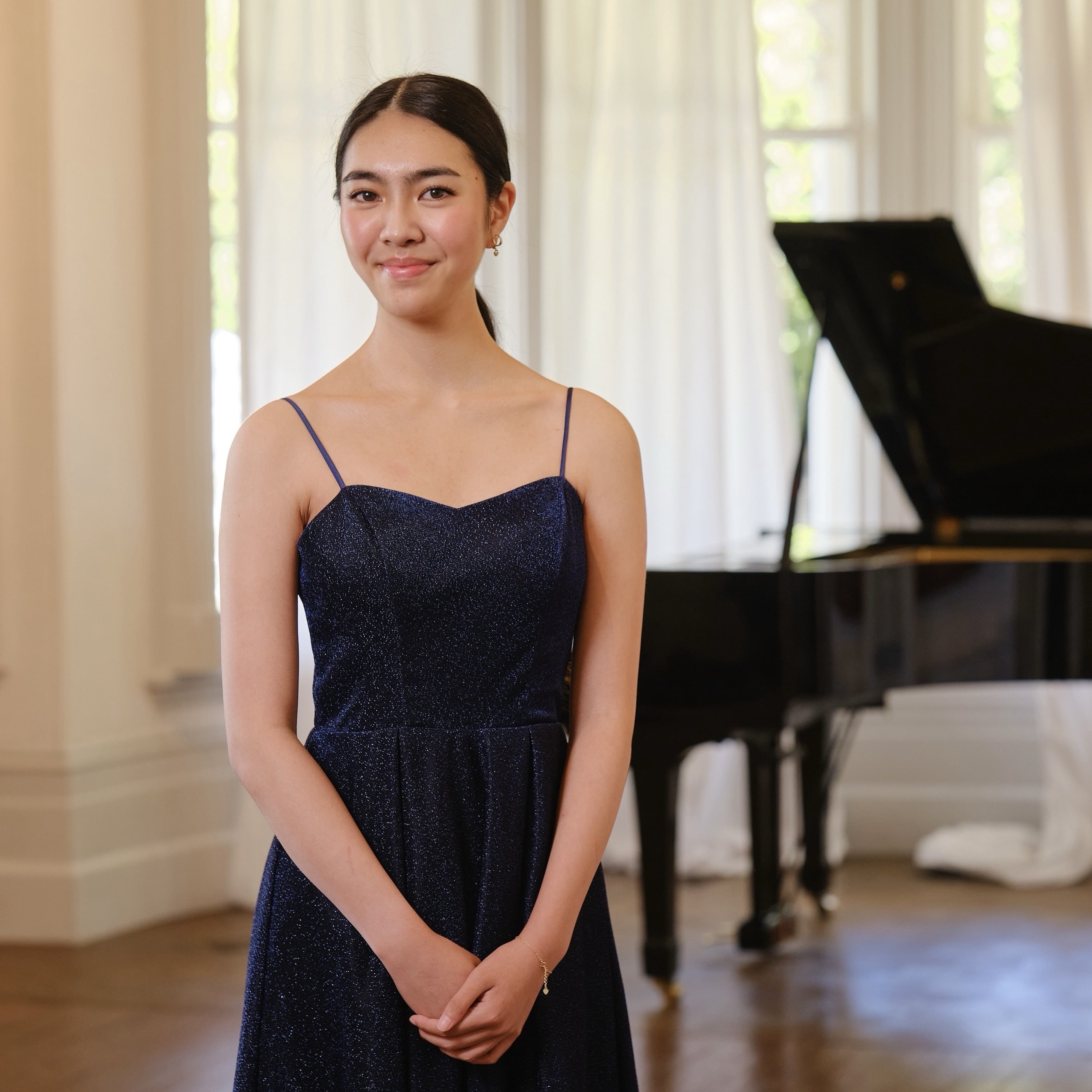 Photo of a young woman of Asian descent wearing a black eveniong gown standing in front of a grand piano.