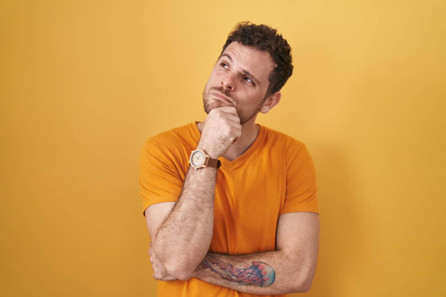A man holds his chin looking thoughtful. He is wearing an orange tee shirt and stands against a yellow background.